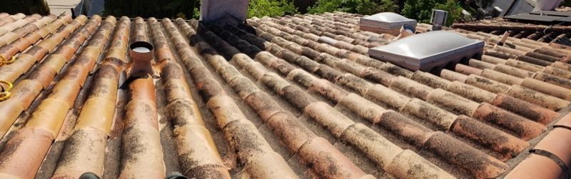 Rusted terracotta roof tiles with a skylight and chimney on a rooftop under bright sun.