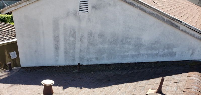 White stucco building side wall with roof and small vent, viewed from above under bright sunlight