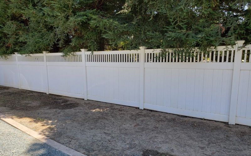 White vinyl fence along a gravel driveway with trees behind it.