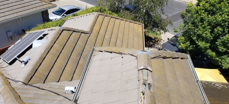 Aerial view of tan roof panels with damage and missing shingles on a house near trees and a driveway