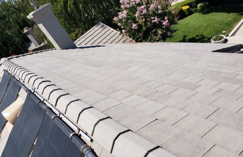 Sloped gray shingled roof with chimney, seen from above beside a green yard and flowering trees
