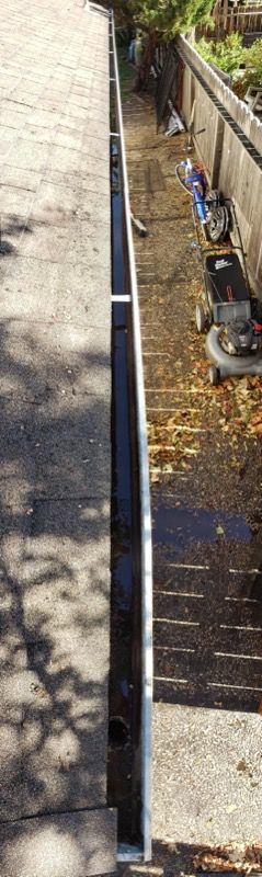 Narrow outdoor walkway beside a wall and fence, with pipes and scattered leaves along the edge.