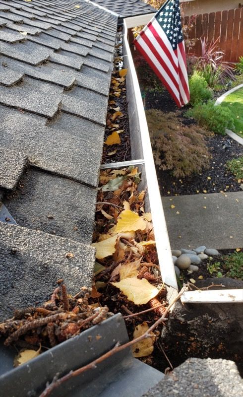 Damaged roof gutter clogged with leaves beside an American flag and garden path