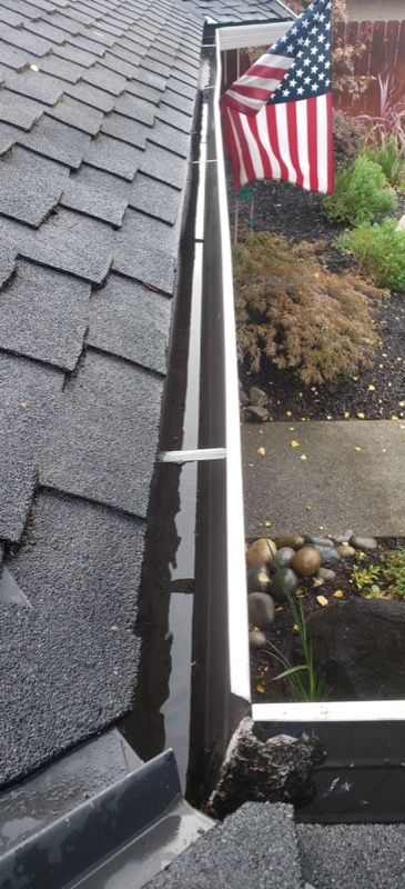 Close-up of roof shingles and a white gutter beside a walkway and American flag in a garden.