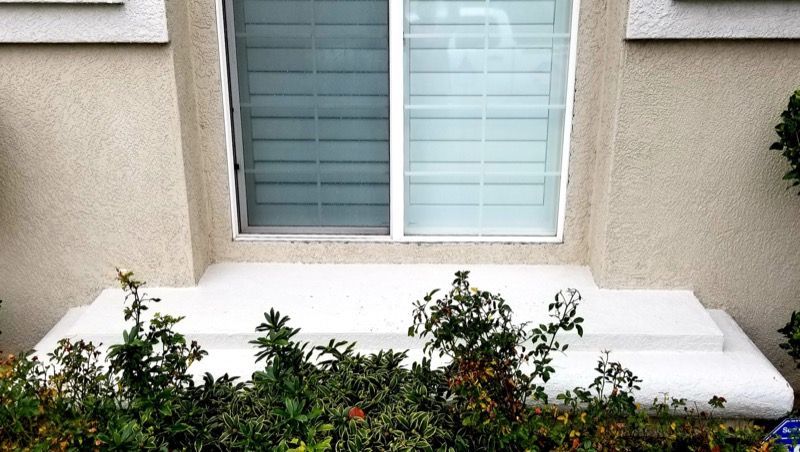 White outdoor bench beneath a window, with shrubs and flowers in front of a stucco wall.