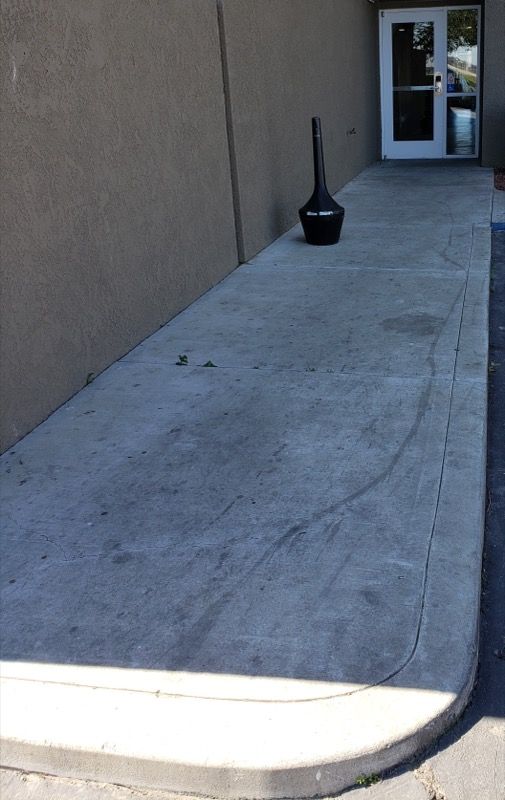 Empty concrete walkway beside a tan wall, with a black trash can near a doorway at the far end.