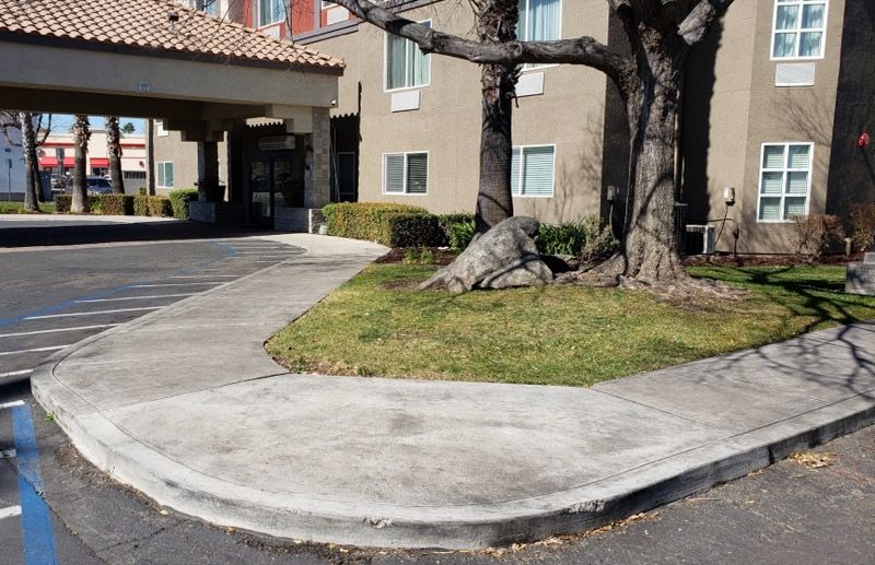 Concrete curb ramp beside a driveway, with a tree and apartment building in the background.