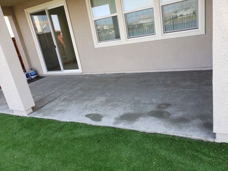 Covered concrete patio with a sliding glass door, windows, and wet stains beside green grass.