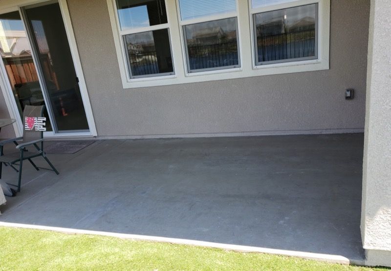 Covered concrete patio beside a house, with windows, sliding door, and a patch of grass in front.