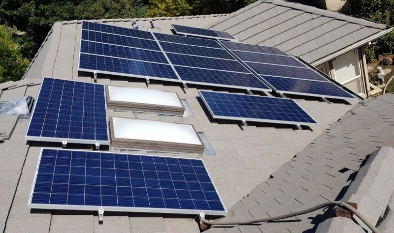 Solar panels mounted on a gray shingled roof around skylights, with trees in the background.