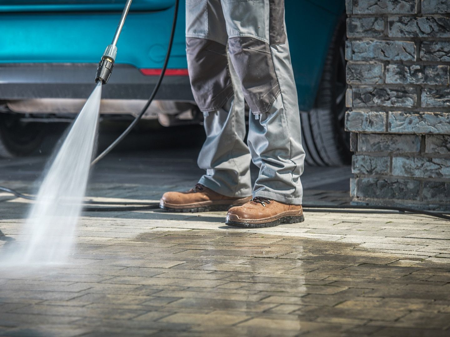 Person pressure-washing a driveway beside a blue vehicle and stone wall