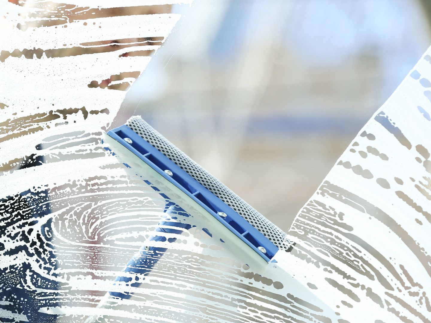 Blue window squeegee cleaning a glass pane with water streaks and soap suds