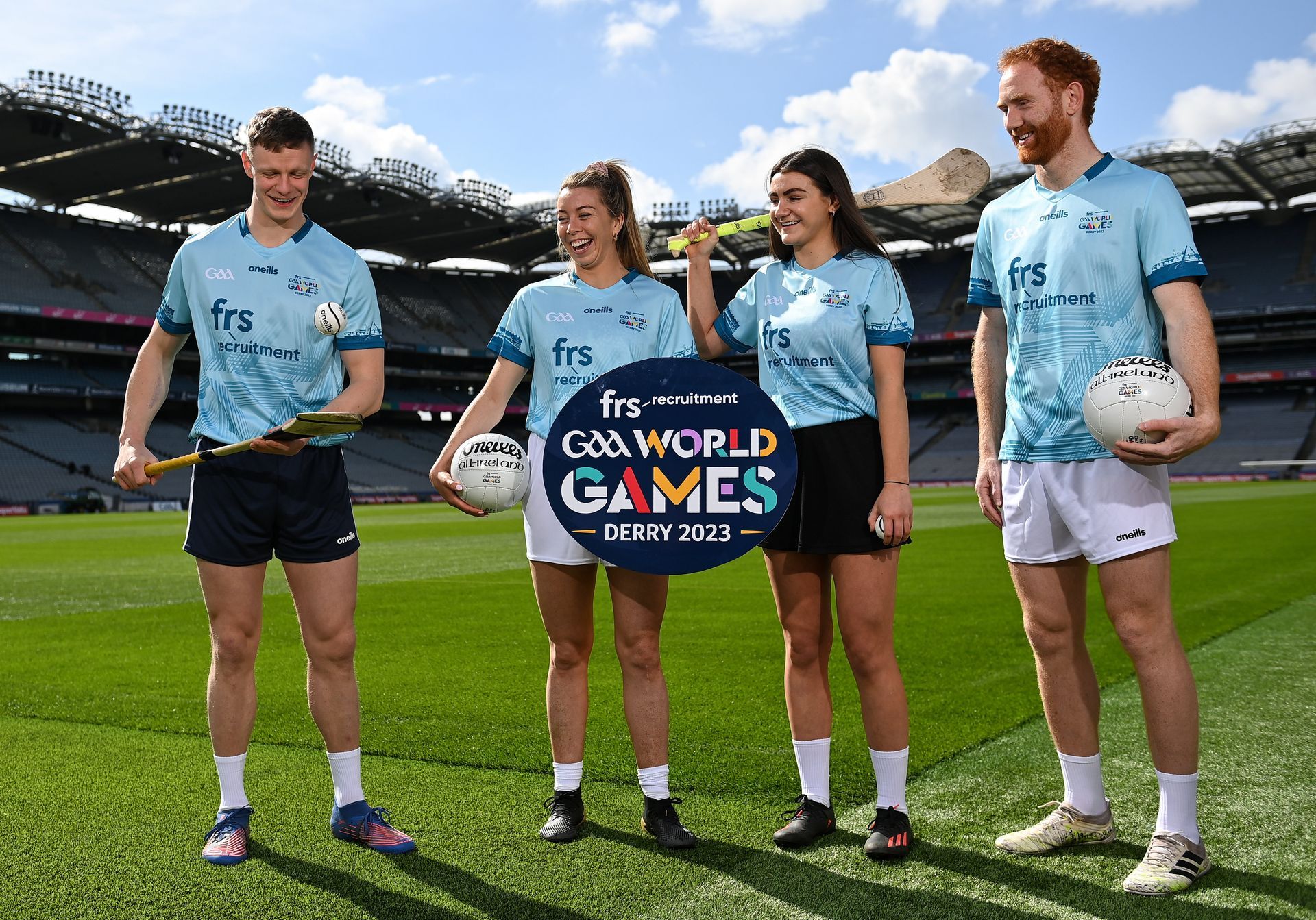 Four people in light blue shirts on a green field holding GAA sports equipment and the event logo.