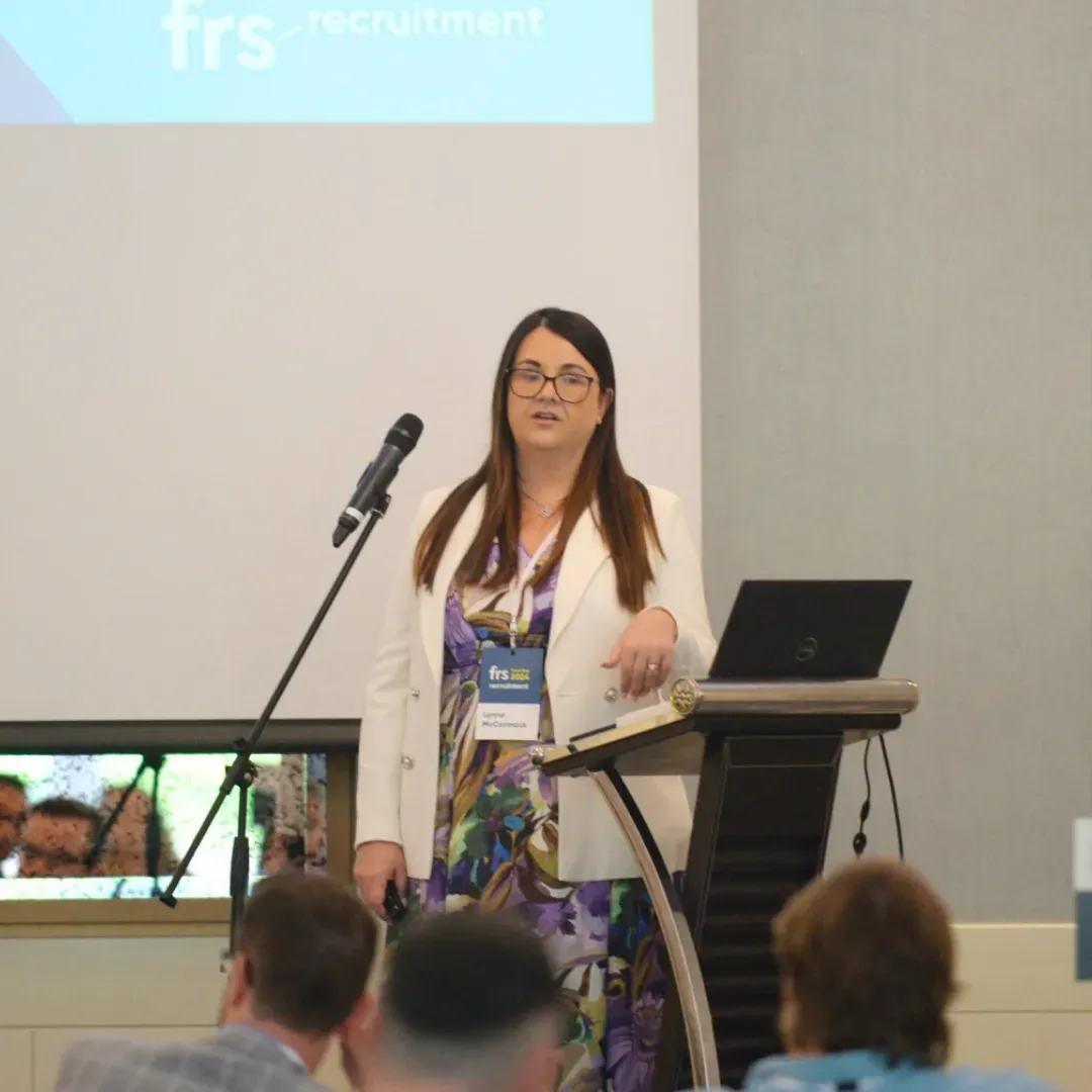 Woman presenting at a conference, wearing a white blazer, standing at a podium.