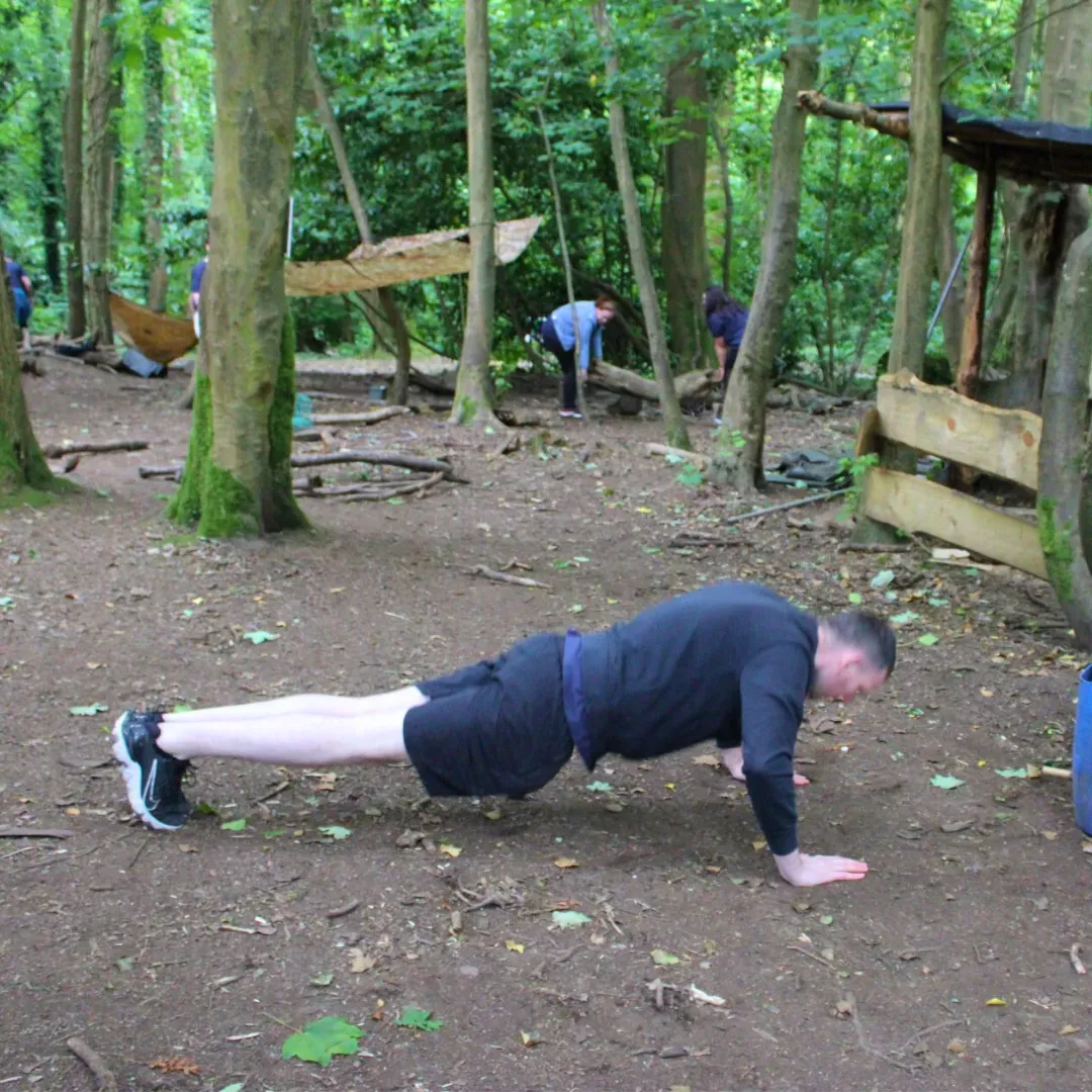 Man doing push-ups on dirt ground in a wooded area. Other people are in the background near wooden structures.