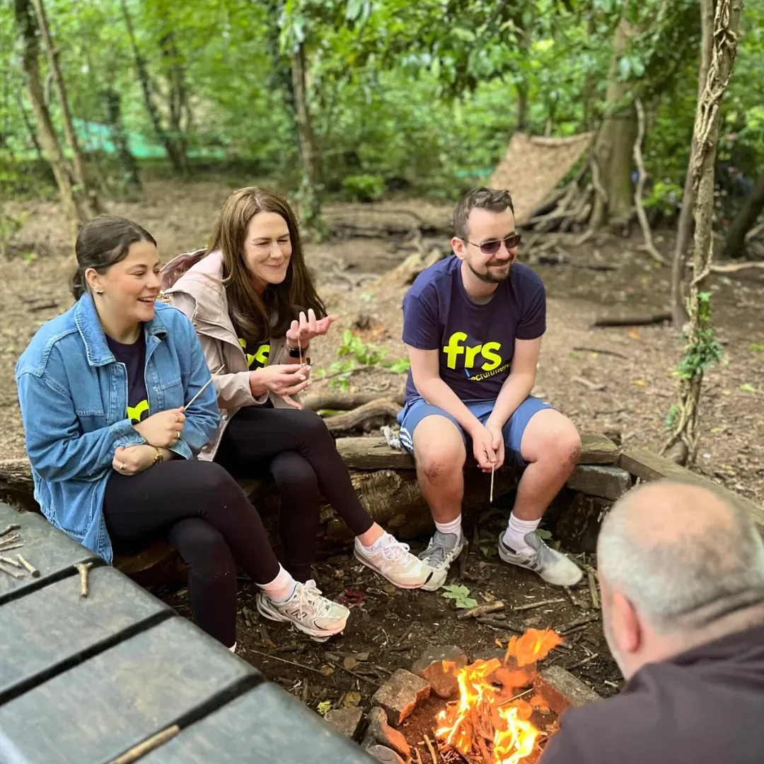 Three people sit by a campfire in a forest, smiling and laughing. A man is partially visible, facing them.