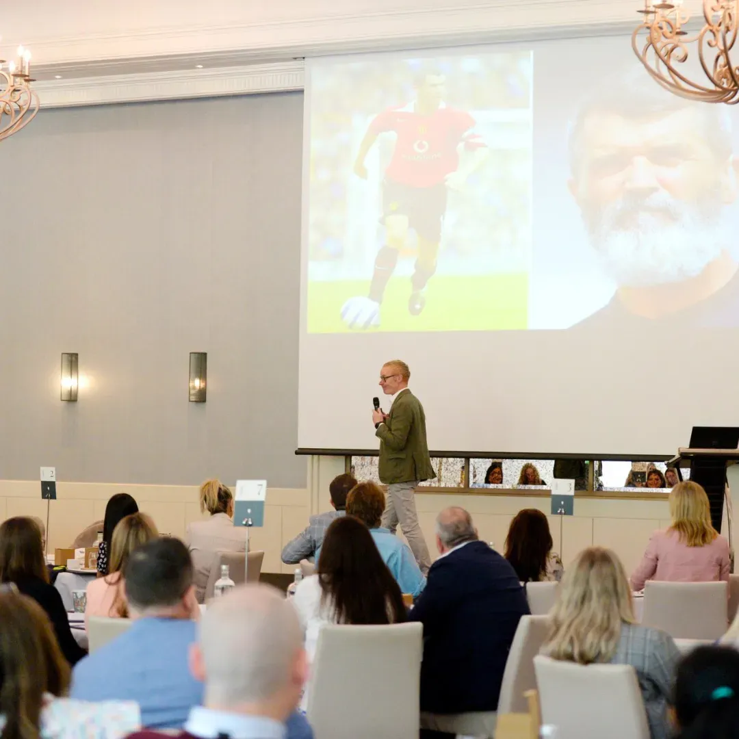 Man giving a presentation to an audience in a conference room with a large screen displaying a soccer player and a portrait.