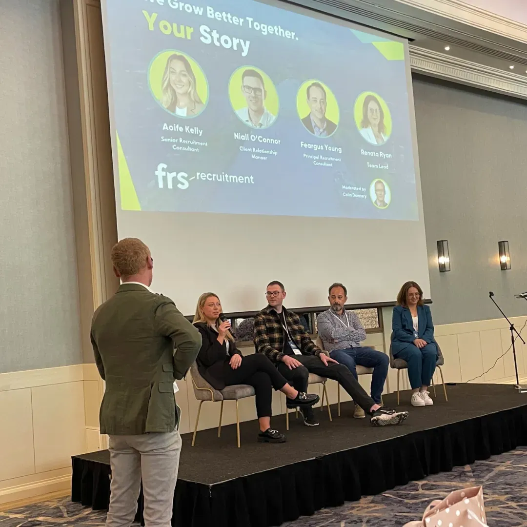Panel discussion at a conference: Speakers on stage with presenter; screen displays speaker photos.