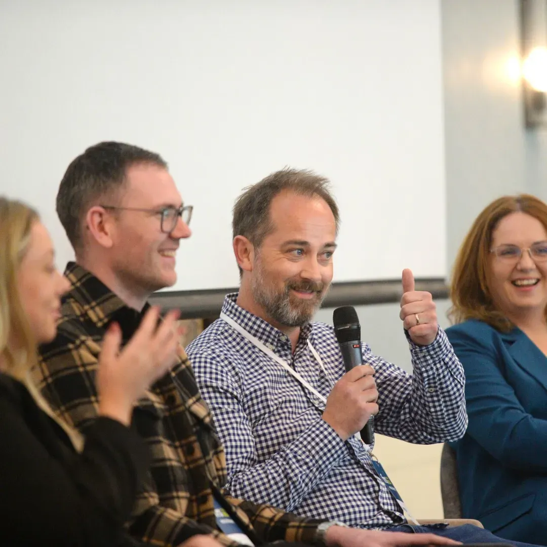 Panel of four people seated, one man giving a thumbs-up while speaking into a microphone. Smiling faces, likely an event.
