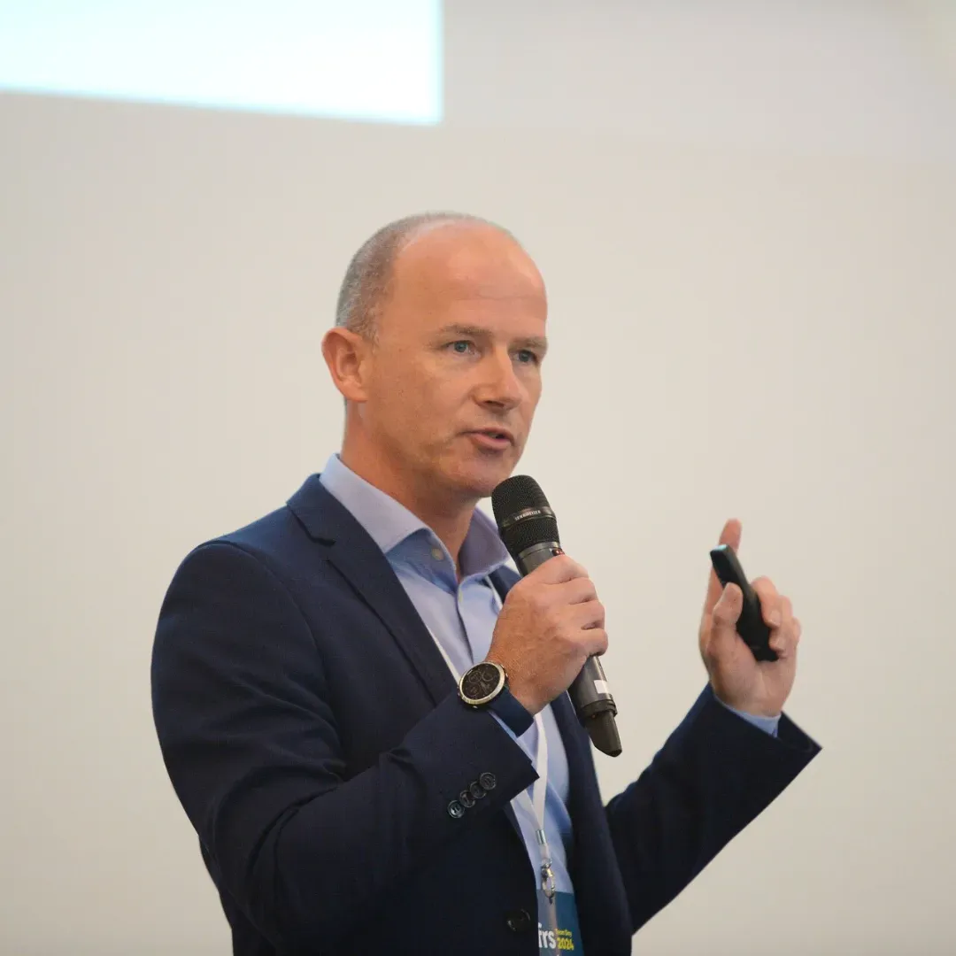 Man in a blue suit speaks into a microphone, gesturing with his hand.