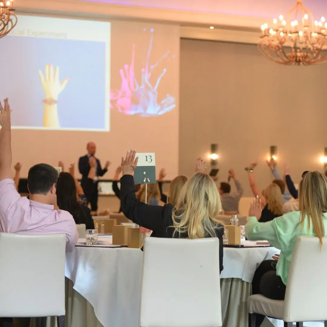People raising hands in a conference room during a presentation; projector screen displays a hand and an abstract image.