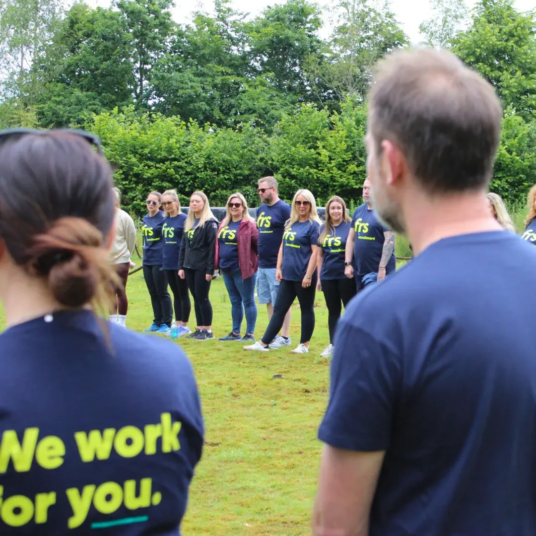 People in matching shirts stand on grass, facing the camera. The shirts say