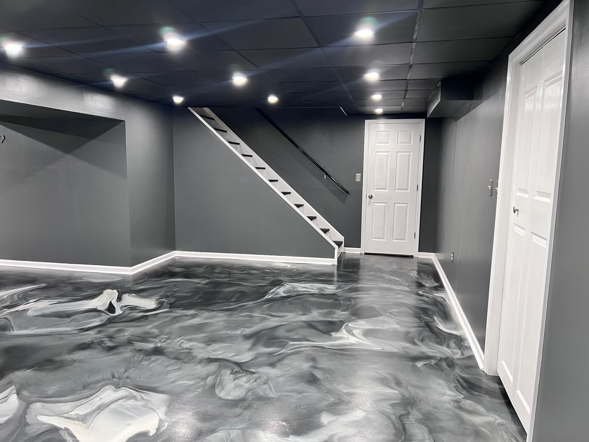 Empty gray basement room with marbled floor, white doors, and a staircase along the back wall