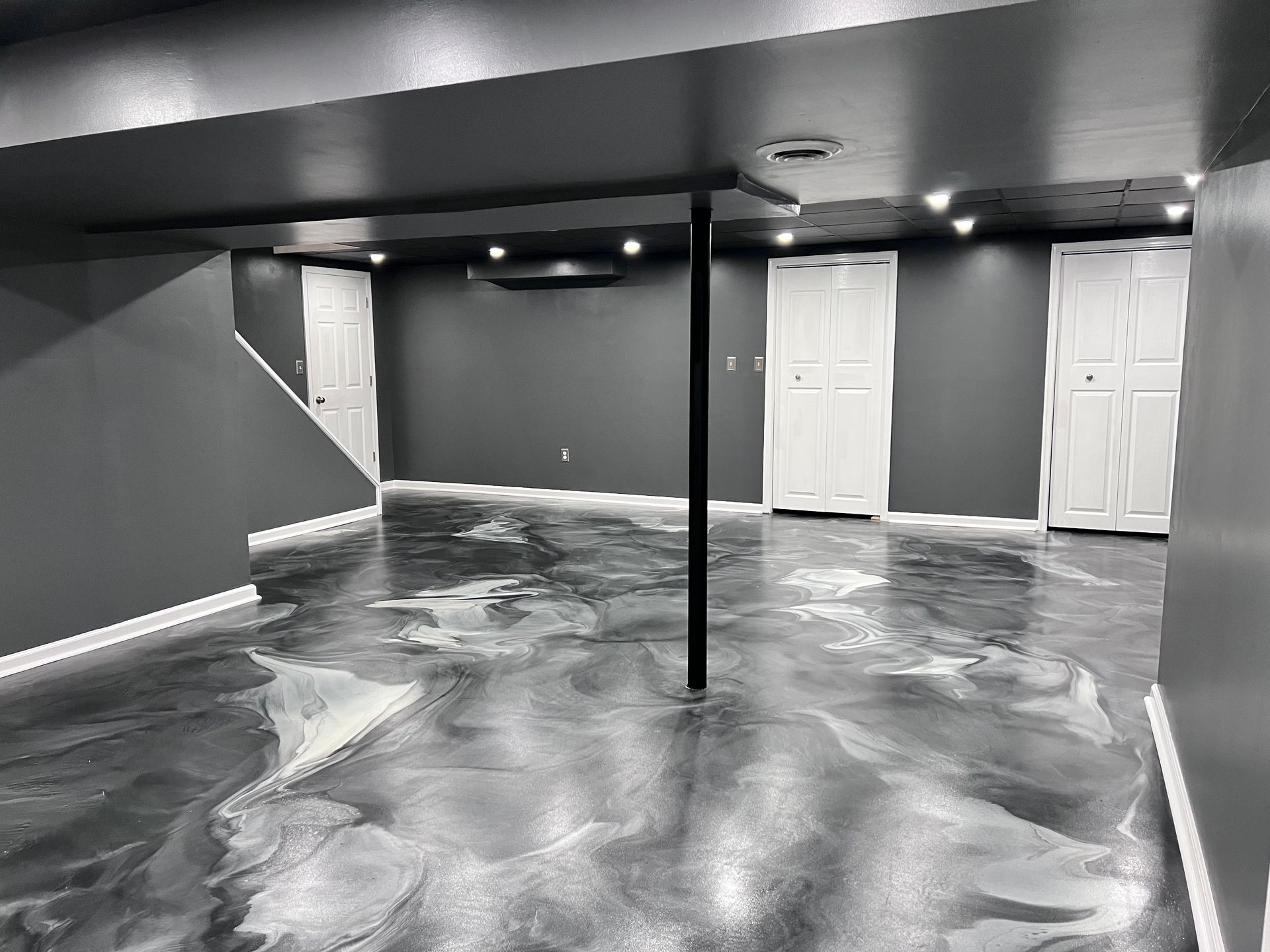 Empty basement room with glossy gray floor, black support pole, and white doors along the back wall