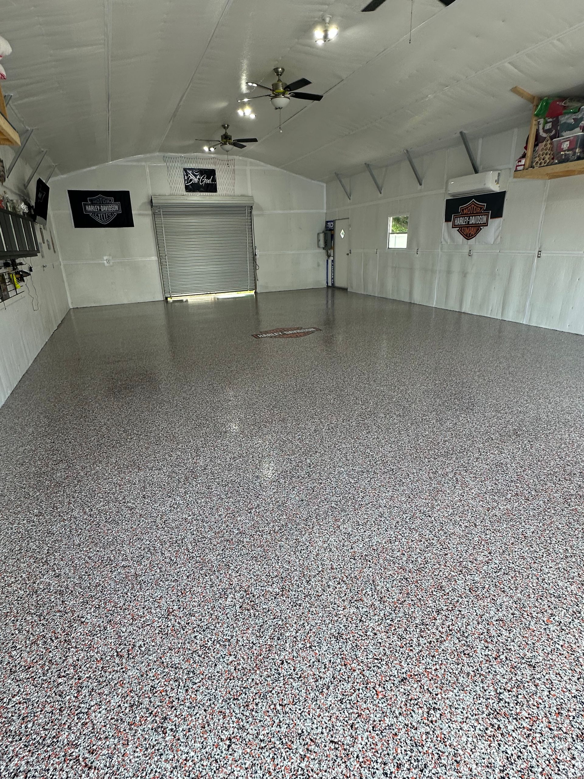 Empty indoor garage with white walls, speckled epoxy floor, ceiling fan, and a roll-up door.