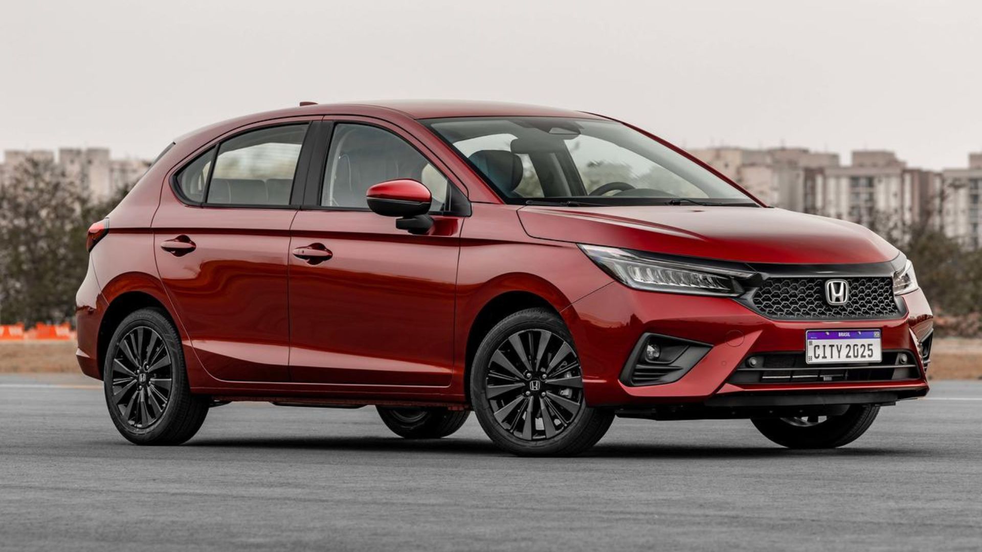 Red Honda hatchback parked on a wet road in an overcast outdoor setting.