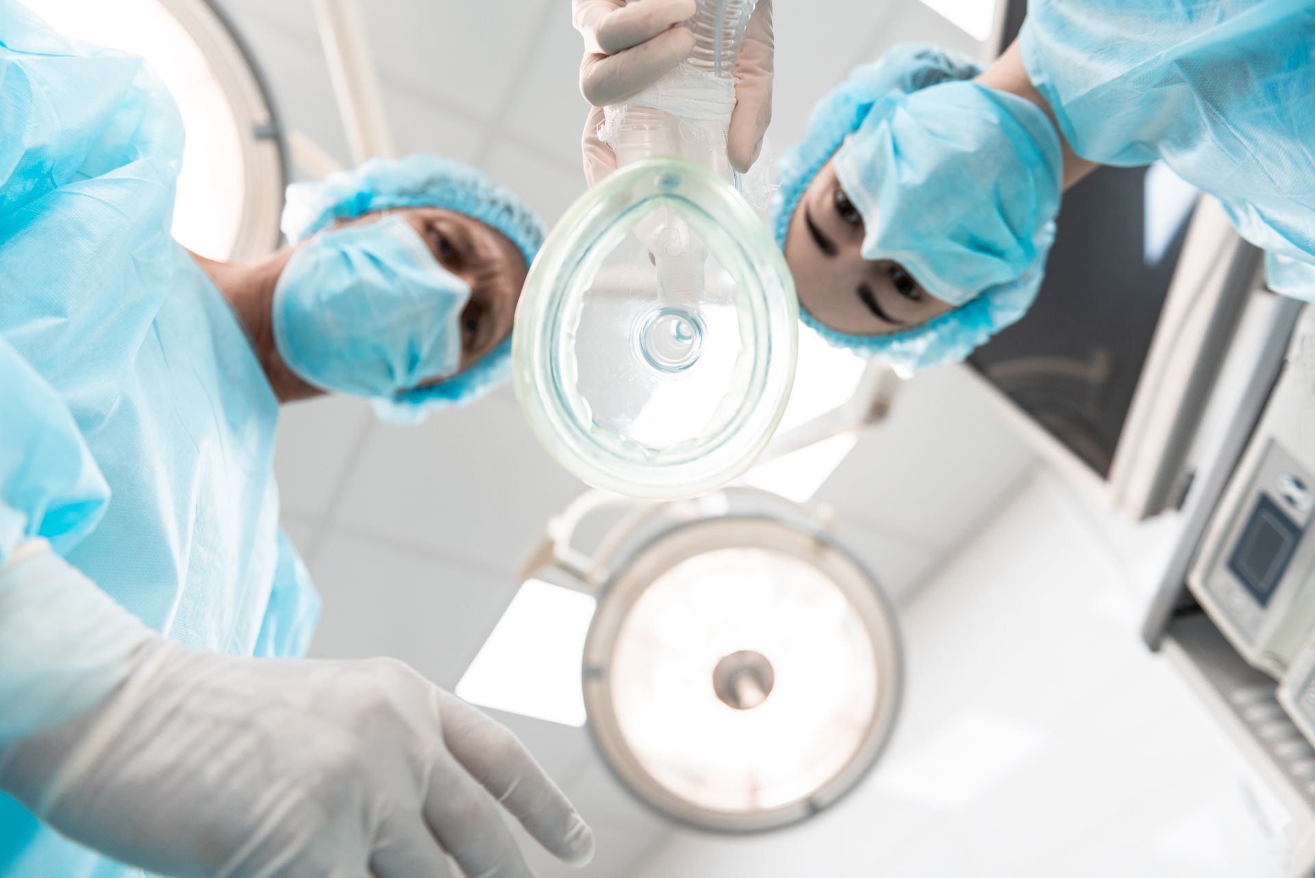 Two surgeons in blue scrubs examining a patient under bright operating room lights apply an anesthesia mask