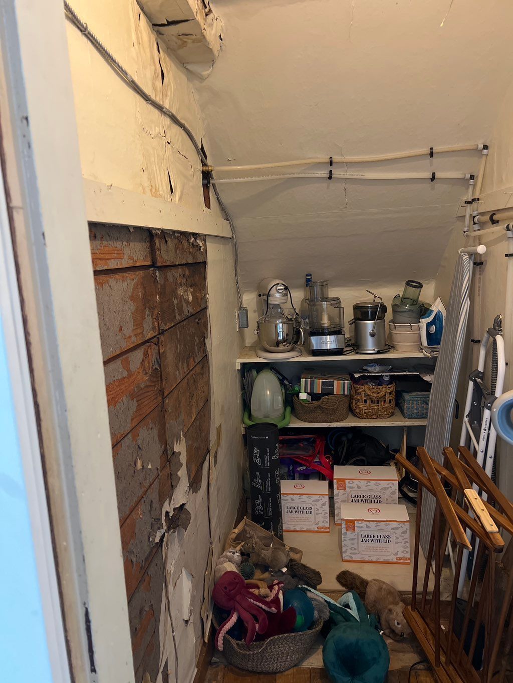 A narrow, cluttered storage closet featuring exposed brick, wooden shelves with kitchen appliances, and boxes on the floor.