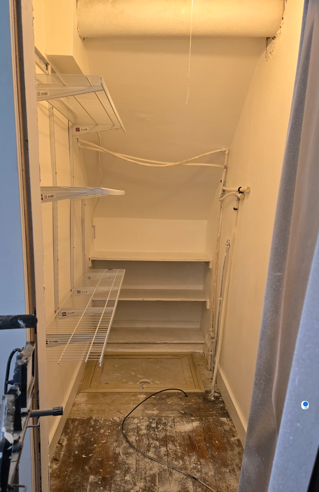 A small, narrow closet with white walls, wire shelving on one side, fixed shelves at the back, and a unfinished floor.