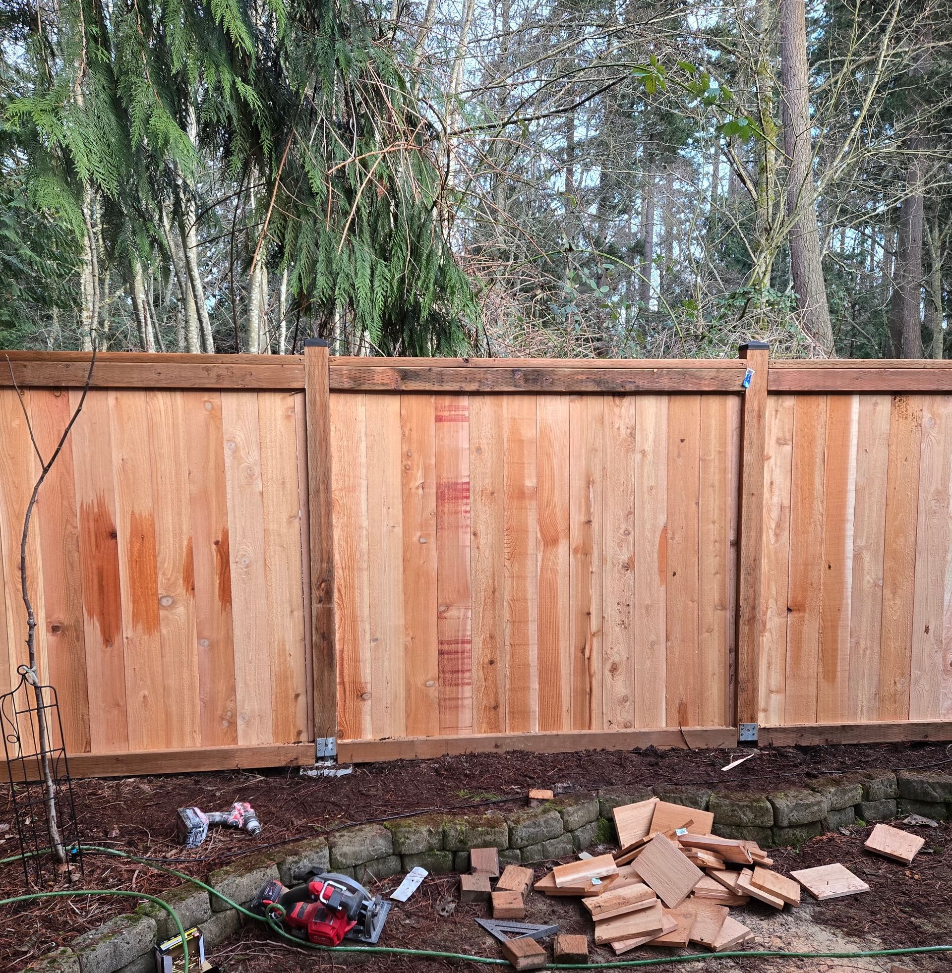 A new cedar wood fence stands in a backyard with wood scraps on the ground in front of a stone garden border.
