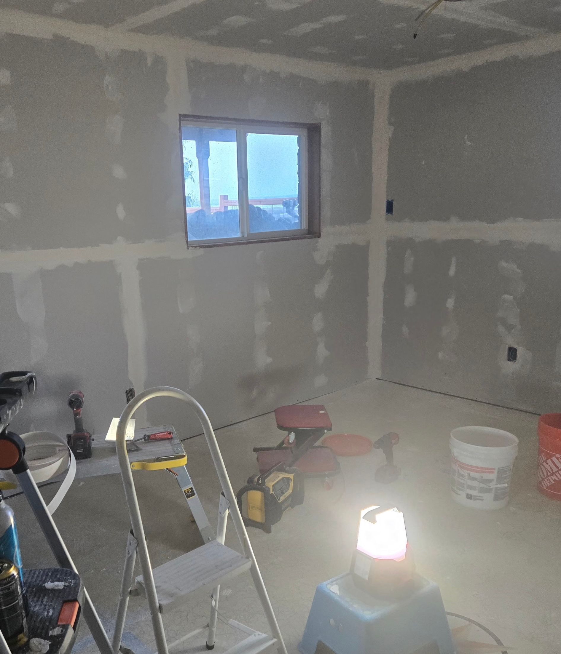 An unfinished room under construction with bare drywall, mudded seams, tools, a ladder, and a bright work light on the floor.