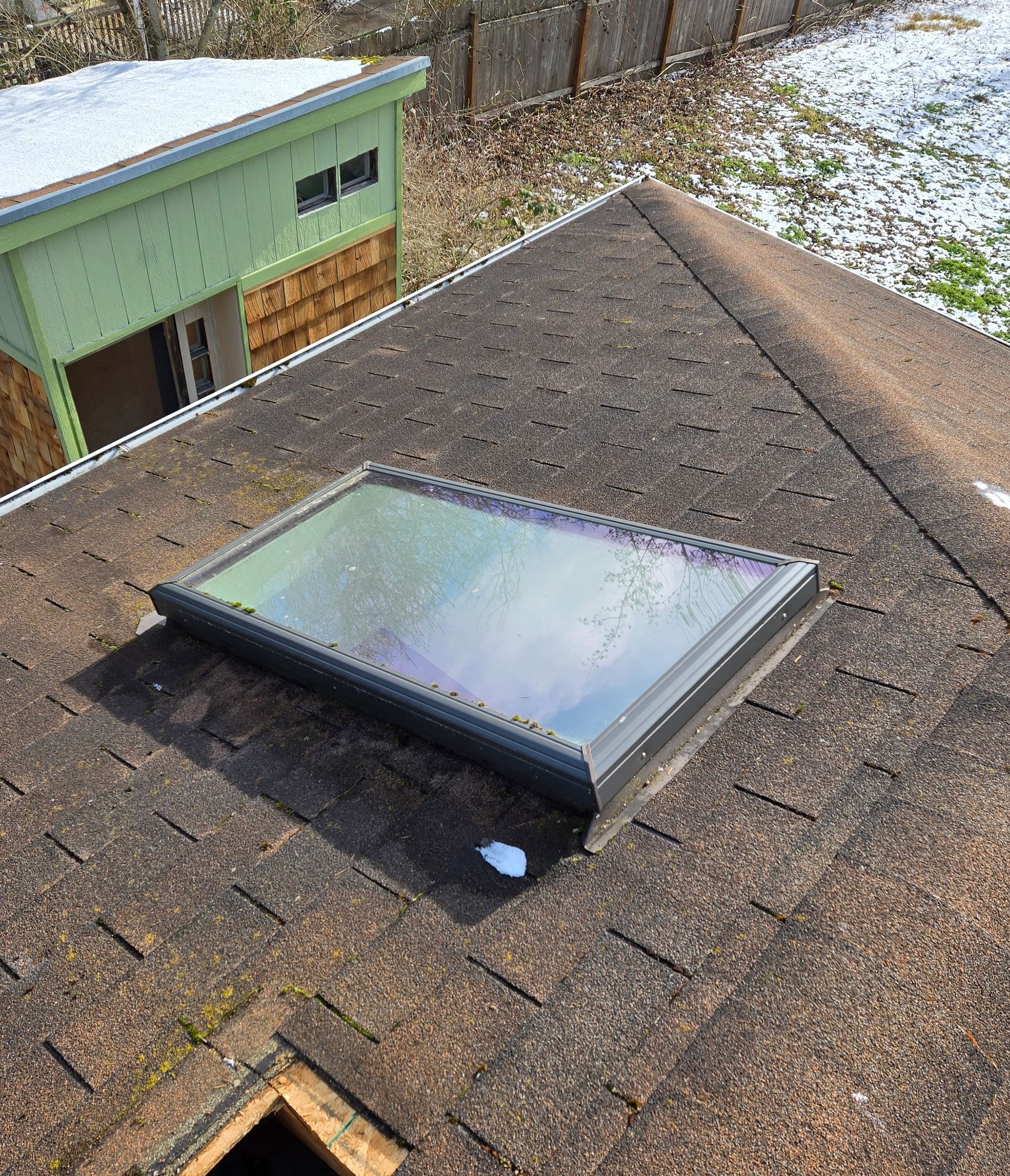 A rectangular skylight installed on a dark shingled roof, with a glimpse of a nearby green building and snowy ground.