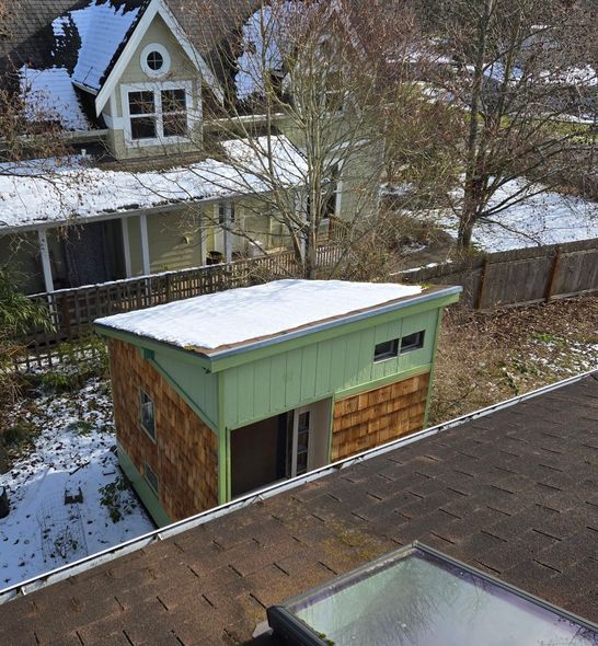 A snow-covered shed with wood siding stands next to a larger house, seen from a high angle near a roof skylight.