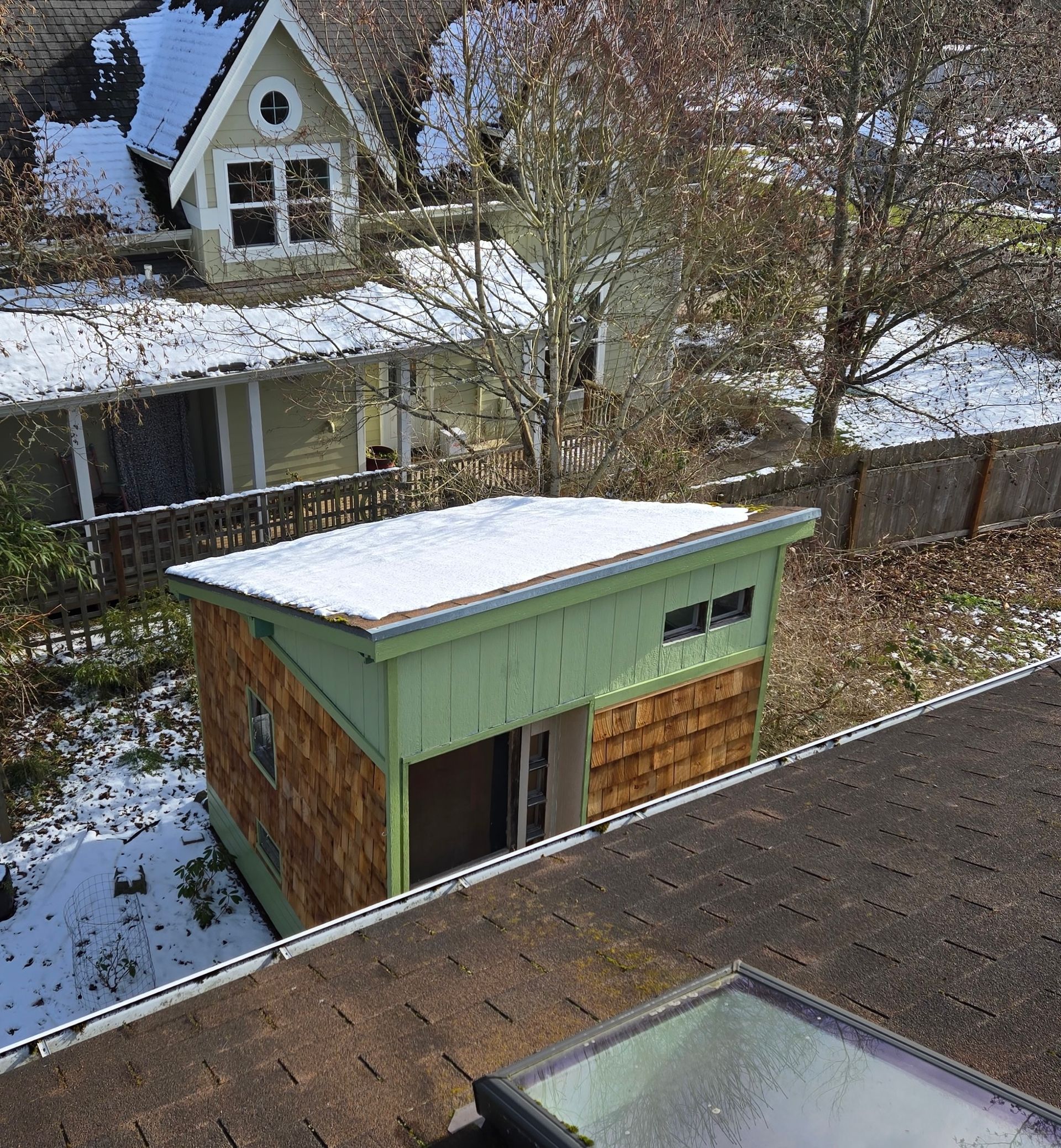 A high-angle view of a shed with a light green exterior and cedar shingles, topped with snow, next to a larger house.
