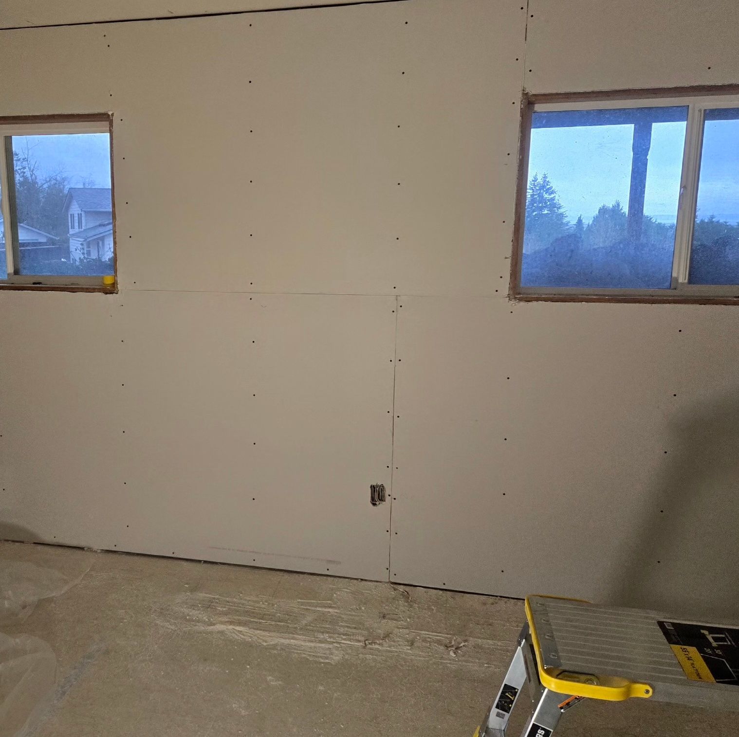 Two windows are centered in a room with unfinished drywall walls and a concrete floor, with a step stool in the corner.