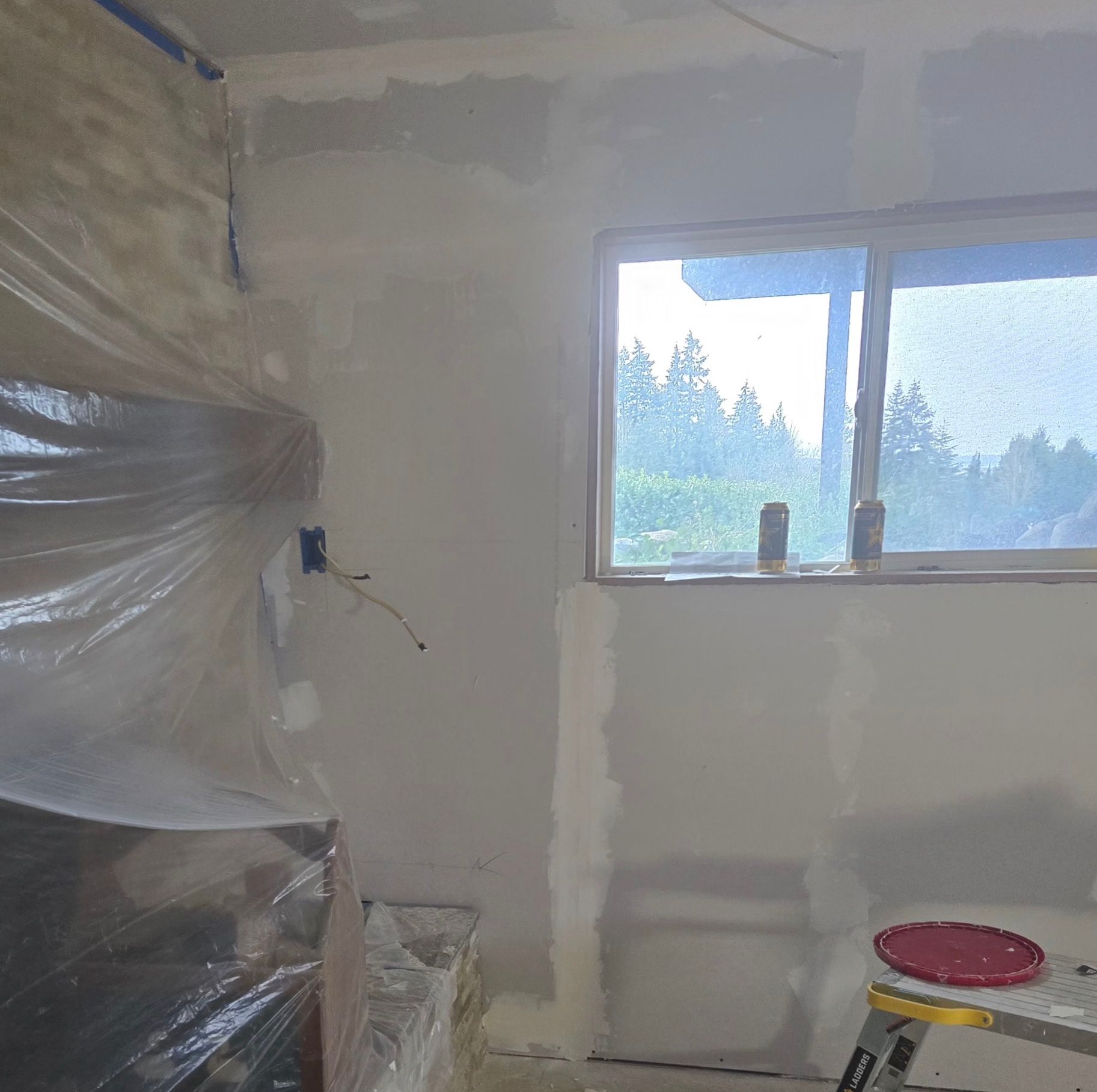 A room undergoing interior renovations with exposed drywall, taped seams, a window, and plastic sheeting covering an area.