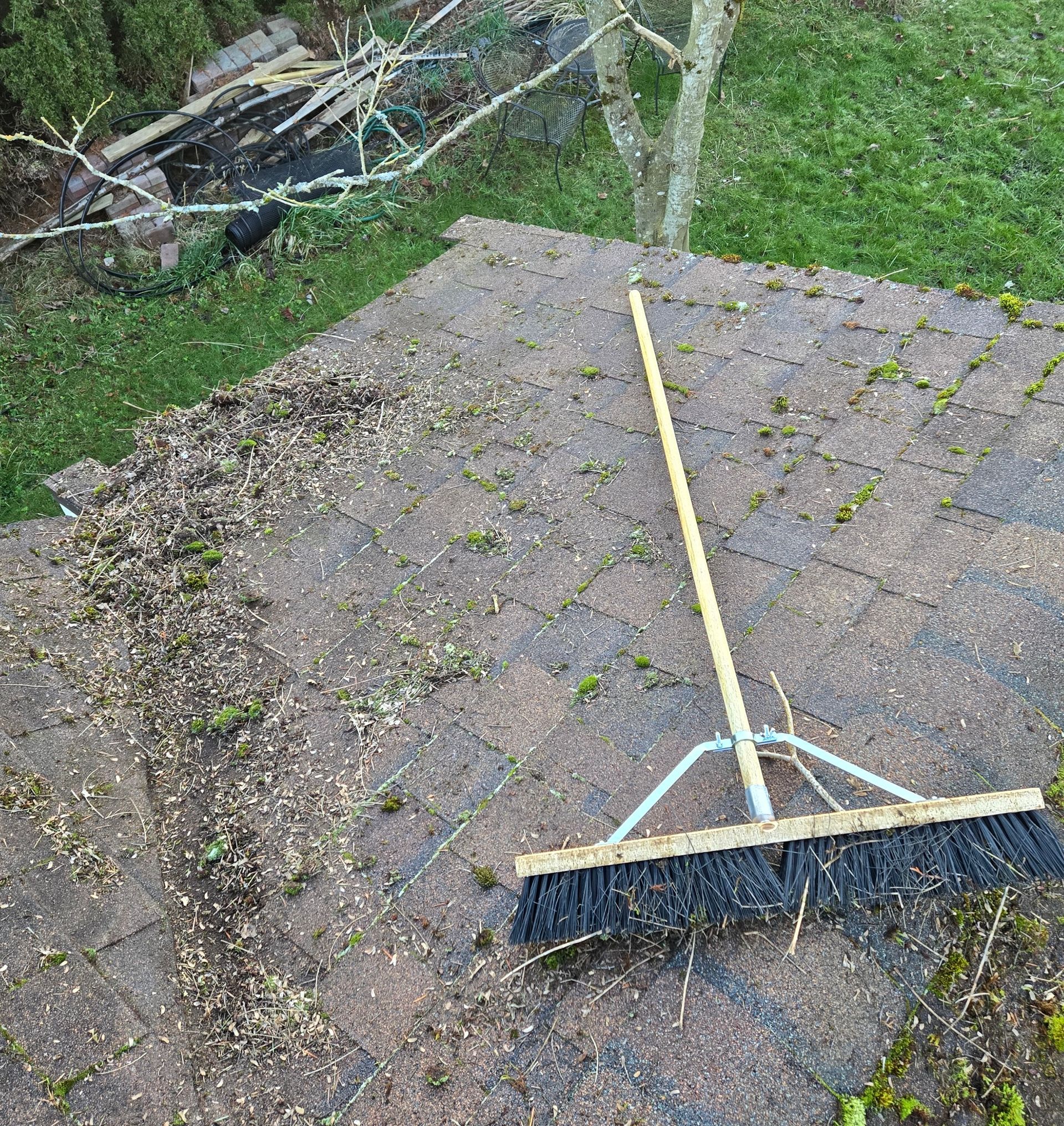 A long-handled push broom sits on a brown brick patio next to a pile of swept-up yard debris.