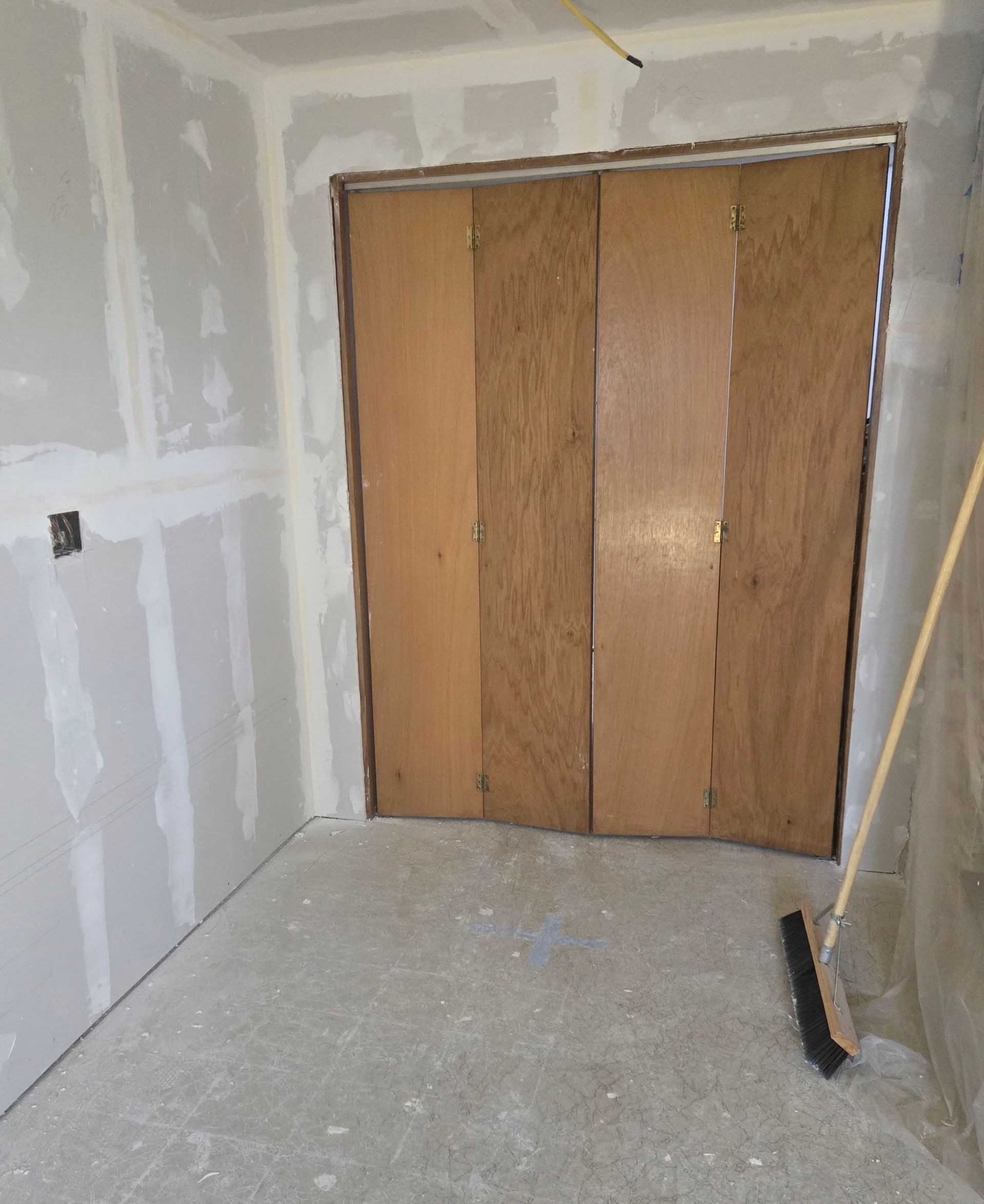 Unfinished room featuring drywall-taped walls, a concrete floor, and a wooden bi-fold door, with a broom in the corner.