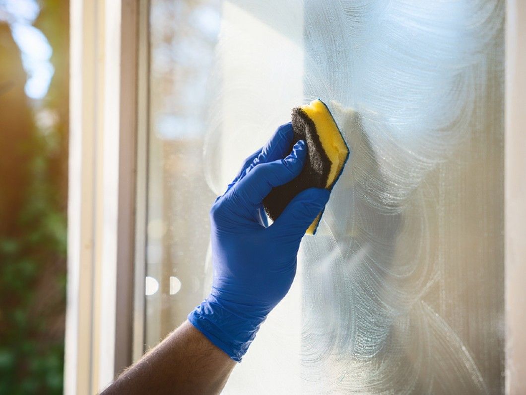 Gloved hand wiping a window with a sponge, leaving streaks; window frame visible.
