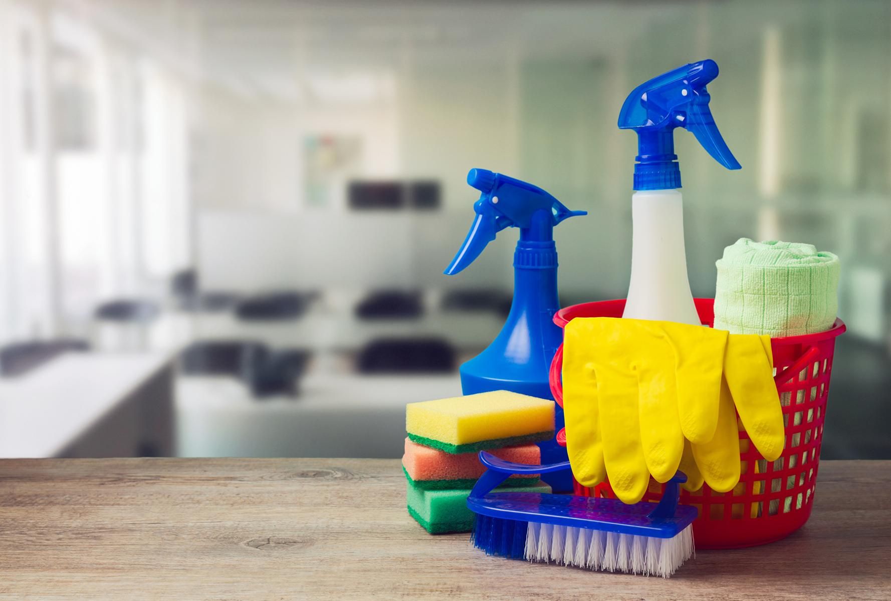 Cleaning supplies: spray bottles, sponges, gloves, and a brush in a red basket on a wooden table.