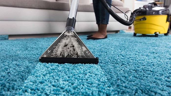 Carpet being cleaned with a vacuum cleaner, turquoise carpet, person's legs visible, yellow vacuum in background.