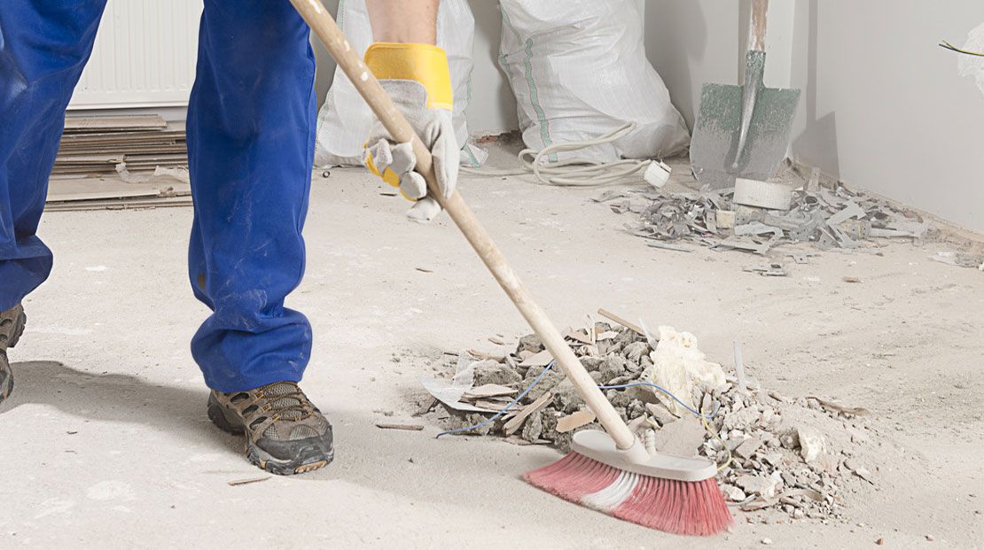 Person in blue coveralls sweeping debris on a concrete floor with a broom.