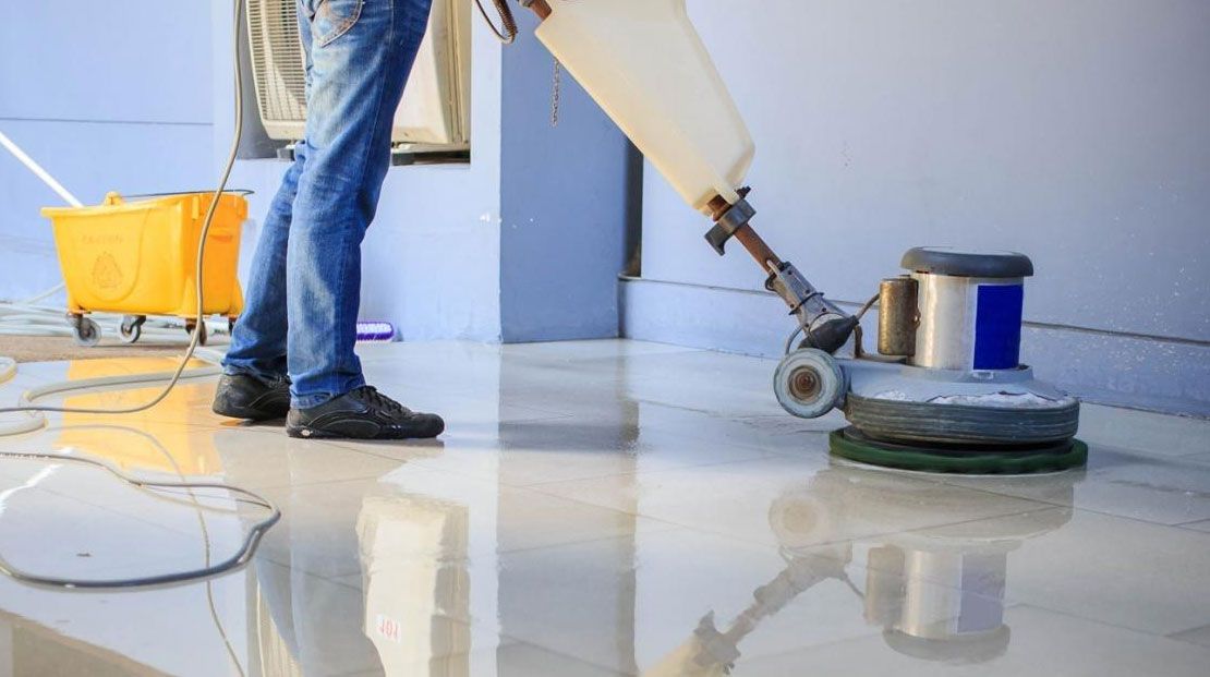 Person using a floor polishing machine on a glossy tile floor, with a yellow bucket nearby.