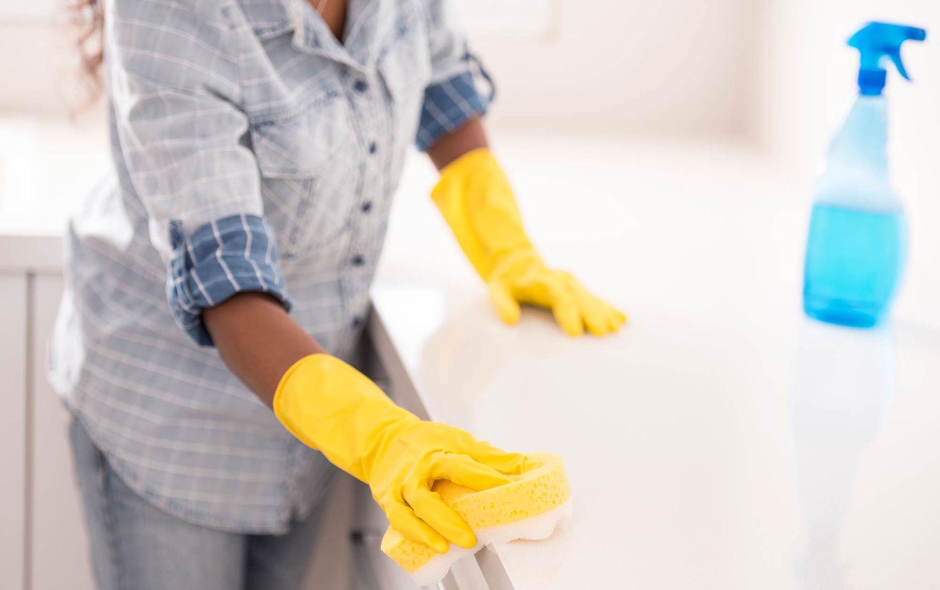 Person wearing yellow gloves cleans a white surface with a sponge, spray bottle in background.