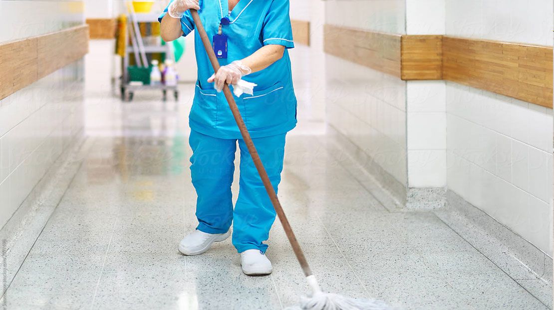 Person in blue scrubs mops a shiny hospital hallway floor. Cleaning cart visible in the background.