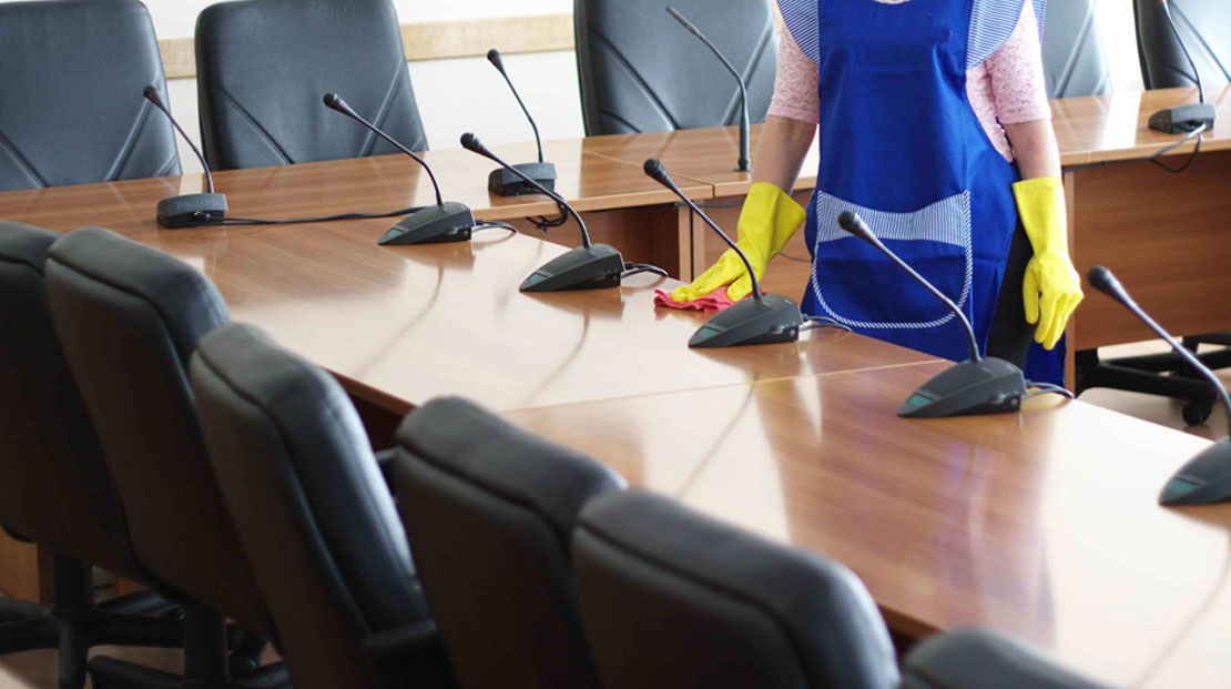 Person in blue apron and yellow gloves cleaning a conference table with microphones.